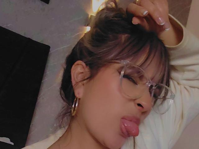 AmiiRosse, la langue tirée, portant des lunettes, des boucles d'oreilles pendantes et une chemise blanche, avec les cheveux tirés par une autre main, dans un selfie légèrement flou..