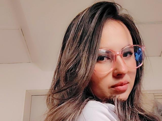 AmiiRosse con occhiali rosa, capelli castani ondulati e labbra piene, che si guarda oltre la spalla, indossa una maglietta bianca, in un selfie..