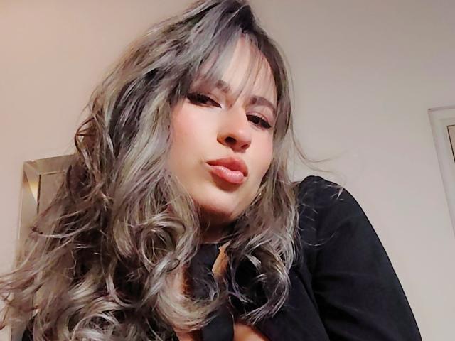 AmiiRosse ha i capelli ondulati, di un colore marrone-grigiastro, con la frangia, labbra piene con un leggero lucido rosa, e indossa una maglietta nera. Guarda in basso con un sorriso leggero e sicuro..