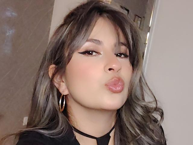 AmiiRosse con labbra piene, eyeliner nero spesso e lunghi capelli ondulati grigio-marrone, che indossa orecchini a cerchio dorati e una collana nera, che guarda verso la fotocamera..