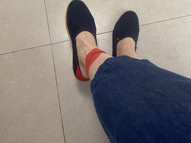 Nalini's legs with blue jeans, black slip-on shoes, and red underwear partially visible, showing a thin ankle with visible veins on a beige tiled floor.