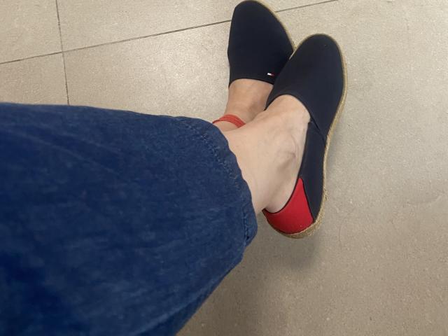 Nalini's legs in dark blue jeans, wearing black flats with red soles, on a tiled floor. Her ankle is slightly visible, showing a red ankle bracelet.