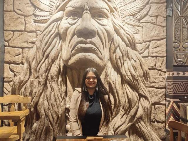 Katherin, com longos cabelos pretos, sorrindo, vestindo uma blusa preta e um casaco bege, está em frente a um grande rosto de leão de pedra, ricamente detalhado. Cadeiras de madeira em ambos os lados..