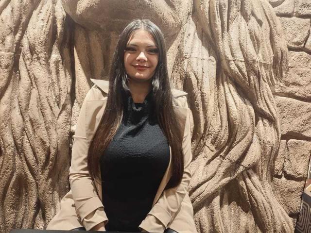 Katherin, con lunghi capelli scuri, una maglietta nera e una giacca beige, sorride, in piedi di fronte a un muro di roccia strutturato, con il suo seno prosperoso e il suo viso carino ben visibili..