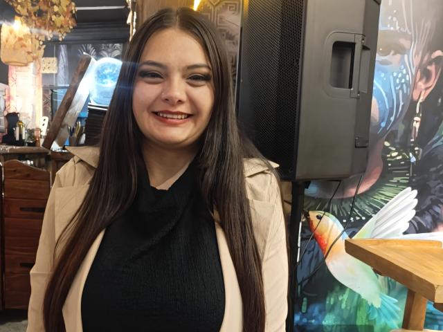 Katherin, une jeune femme aux longs cheveux bruns, souriante, portant un haut noir et une veste beige, se tient devant une télévision avec une illustration d'un oiseau, et met en valeur sa poitrine..