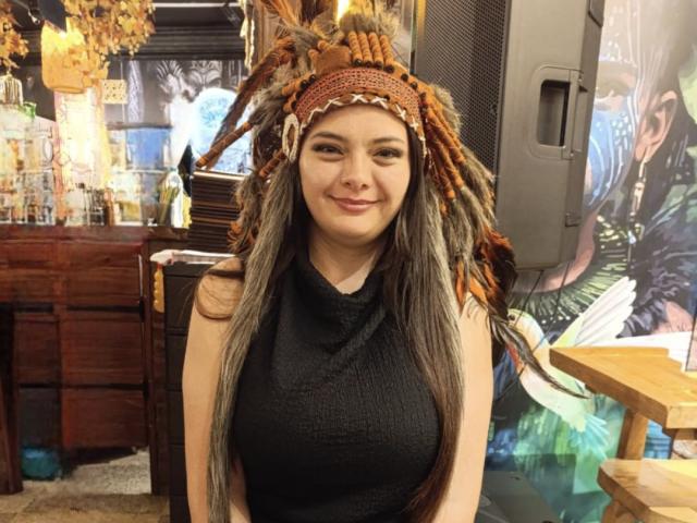 KatherinLatin with long brown hair, black sleeveless top, and orange-brown beaded headpiece, smiling at the camera, showing her tits, standing in a shop with wooden furniture.