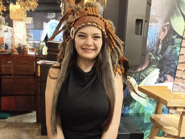 KatherinLatin with long dreadlocks wearing a brown headpiece, smiling, black sleeveless top, showing her tits, standing in a room with wooden furniture and a wall with colorful art.
