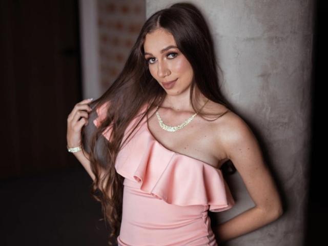 Maittee, con lunghi capelli castani, un abito rosa con una spalla scoperta e una collana dorata, si tiene i capelli, è in piedi contro una colonna grigia, sorride alla fotocamera, mostrando il seno..