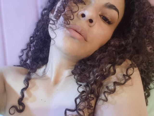 MissSunnyShine with curly dark brown hair, light brown skin, and slightly parted pink lips, lying down, showing her bare shoulders and nose piercing, with a subtle smile.