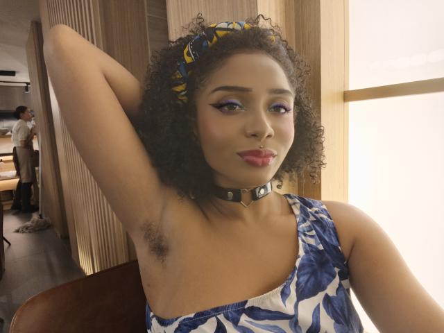 MeganWlliams with curly hair, wearing a blue floral dress and black choker, arm raised to show armpit hair, purple eyeliner, red lipstick, and yellow headband, smiling confidently.