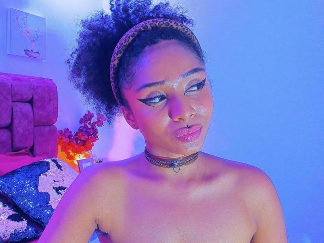 MeganWlliams with curly black hair in a leopard print headband, wearing a choker, looking to the side with a slight pout, topless, blue and purple lighting on her skin.