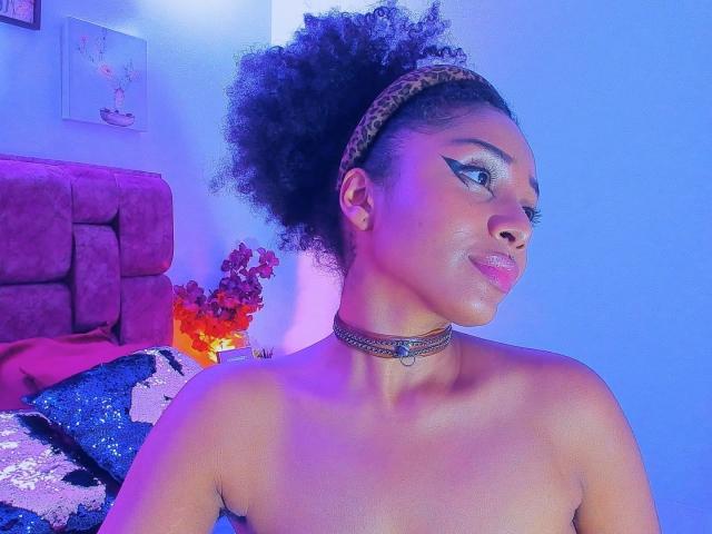 MeganWilliams with a big afro, wearing a leopard headband, choker, and no top, has bold eyeliner, pink lipstick, and a confident look, with blue and purple lighting.