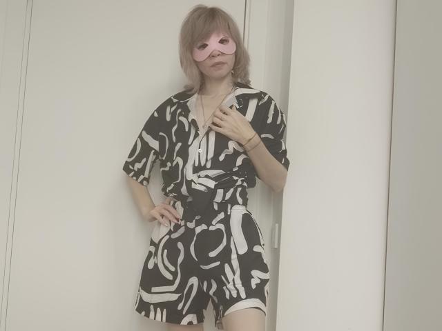 MaryPele with pink mask, wearing black and white abstract pattern dress, standing with hand on hip, light brown hair, white door behind. Boobs not visible, ass not visible.