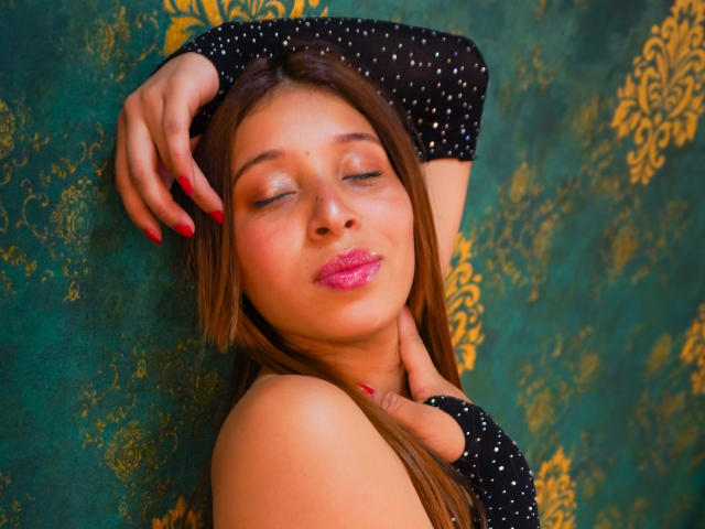 JaneKetty with her eyes closed, wearing a black polka dot top, has her arms above her head, red nails, pink lipstick, and brown hair against a green patterned wall.