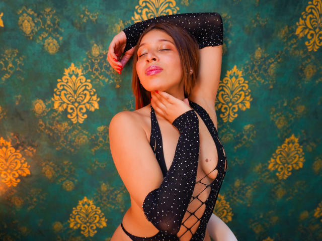 JaneKetty in a sheer black polka-dot bodysuit with lace-up front, eyes closed, arms raised, pink lips, brown hair, and a visible nipple on her right breast.