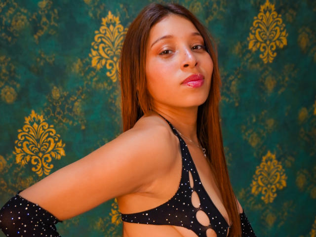 JaneKetty with long brown hair, wearing a black, cut-out top, showing her small tits, pink lips, and confident smile, against a green patterned background.