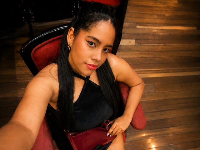 Sienna Taylor mit langen, schwarzen Haaren, die ein schwarzes Top ohne Ärmel trägt, sitzt auf einem roten Stuhl, hält eine rote Handtasche, Holzboden, macht ein Selfie, schaut nach oben, mit pinkem Lippenstift..