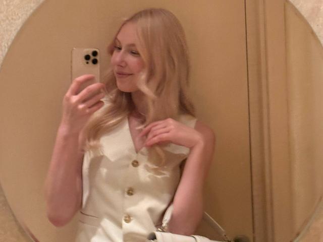 Laurinda Lupacchino, bionda, sorridente, indossa un abito bianco con bottoni, tiene in mano il telefono, sta scattando un selfie, riflesso nello specchio, capelli ondulati, sorriso dolce, pelle chiara, bottoni dorati, casual, sicura di sé, elegante..