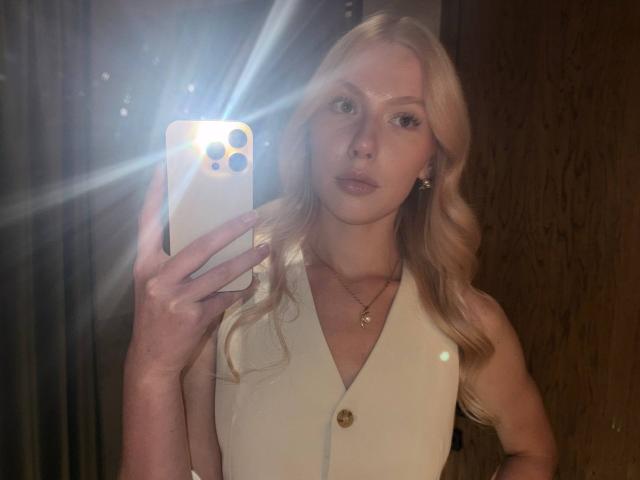 Laurinda Lupacchino mit blonden, welligen Haaren, trägt ein weißes, ärmelloses Kleid mit einem Knopf, hält ein Telefon, macht ein Selfie im Spiegel, leichte Lichtreflexionen vom Telefon..