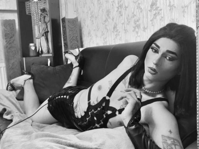 Fotografia in bianco e nero di IrisIrs con lunghi capelli scuri, tatuaggi e una collana di perle, sdraiata su un divano in abbigliamento intimo, con il seno scoperto, e una mano sul petto..
