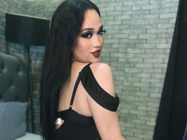 AmaraHugeCock with long black hair, dark makeup, and red lipstick, looking over her shoulder, wearing a black dress with one strap down, showing her back and tits.