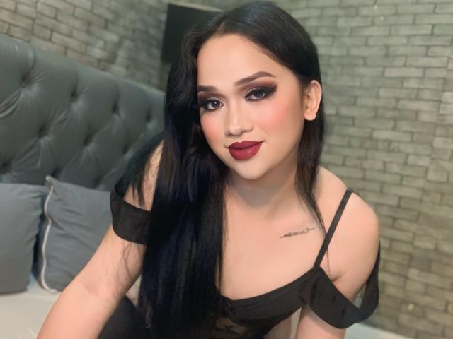 Amara mit langen schwarzen Haaren, rotem Lippenstift und dunklem Augen-Make-up, trägt ein schwarzes Top mit einem Schulterauschnitt und zeigt ein kleines Tattoo auf ihrer Brust. Brüste, Hintern und Brüste sind sichtbar..