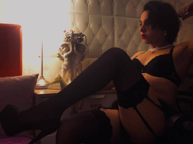 FemCupid vestida con lencería negra, con ligas, medias y tacones, sentada con las piernas levantadas, mostrando sus pechos y glúteos, llevando un collar de perlas, en una habitación con poca luz..