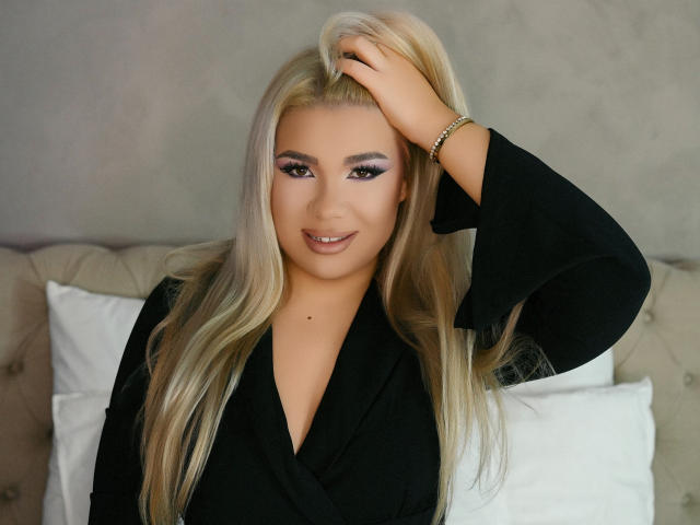 KendalReese, con lunghi capelli biondi, vestito nero e trucco viola sugli occhi, con una mano tra i capelli, sorridente, pelle abbronzata, cuscini bianchi sullo sfondo, braccialetto dorato, sexy, sicura di sé, carina..