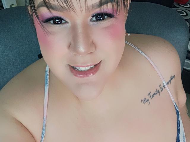 GiiannaMorrys with pink eyeshadow, pink blush, and a nose piercing, smiling at the camera. She has a tattoo on her right shoulder and short brown hair. Her tits are visible.