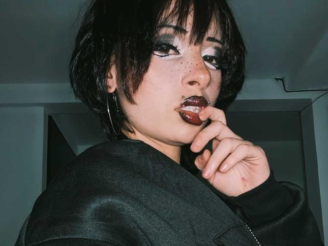 ViolettClay ha i capelli neri e corti, rossetto scuro e un piercing al naso. Si sta mordendo un dito, indossa una giacca nera, con un trucco occhi smokey e delle lentiggini..