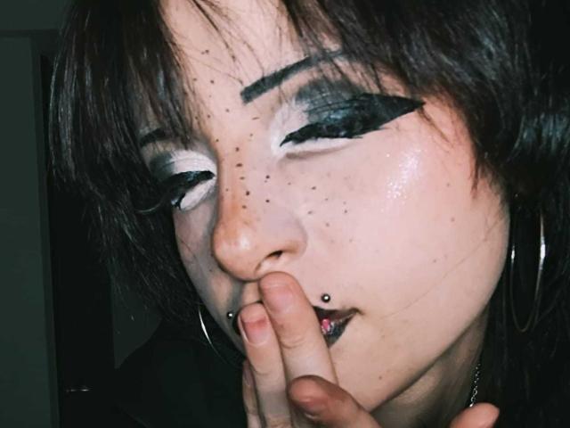 ViolettClay ha un trucco scuro, con un eyeliner nero spesso, ombretto bianco, rossetto nero, lentiggini, capelli neri e un piercing al naso. Si copre la bocca con un dito..