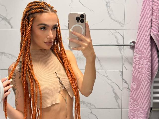 MarissaFex mit orangefarbenen Zöpfen, trägt ein hellbeiges, kurzes, geripptes Oberteil, hält ein Telefon, zeigt ein Tattoo auf ihrem Arm, in einem Badezimmer, mit einem pinkfarbenen Handtuch..