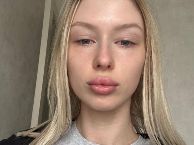 HeidiSmialek's face, showing pale skin, blonde straight hair, light green eyes, and pinkish lips, wearing a gray shirt, with a neutral expression. Boobs not visible.