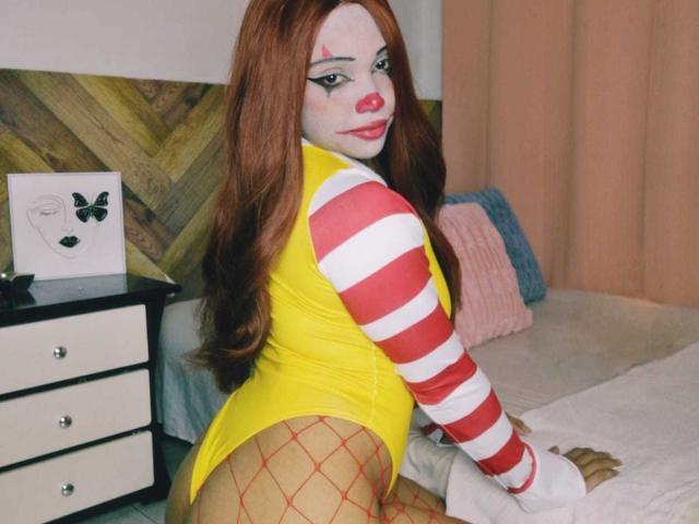 NashaLoule in a yellow bodysuit, red fishnet stockings, and white-red striped sleeves, with painted white face and red lipstick, showing her ass, long red hair, and painted eyebrows.