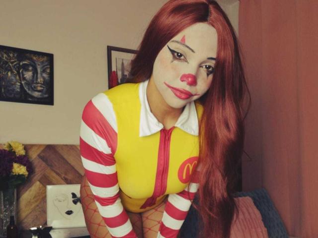 NashaLoule with long red hair, white face paint, red nose, and yellow striped shirt, leaning forward, showing her tits in a red and white top, in a cozy room.