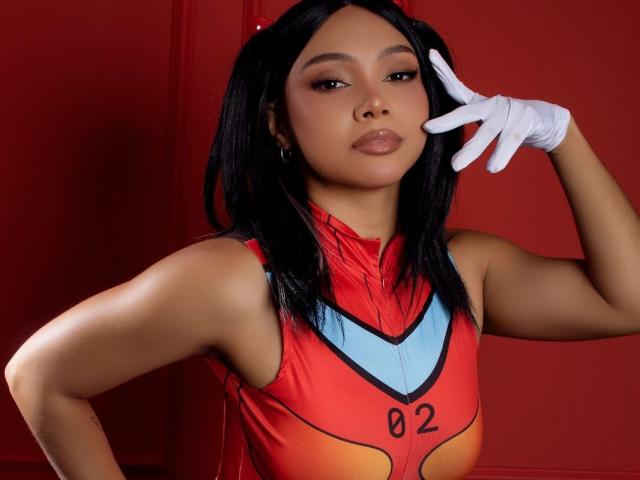 NashaLoule, with dark hair and brown skin, wearing a red and blue superhero costume, white glove, and looking confident, has her hand near her face. Boobs visible under the costume.