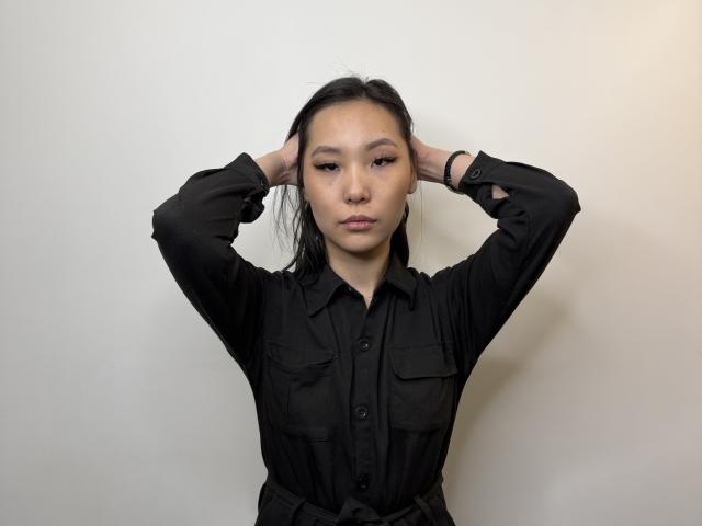 PuraShurts with arms up, black shirt, medium tits, Asian, long black hair, no makeup, serious face, white wall, black bracelet, hands in hair, subtle eyeliner, soft lighting.