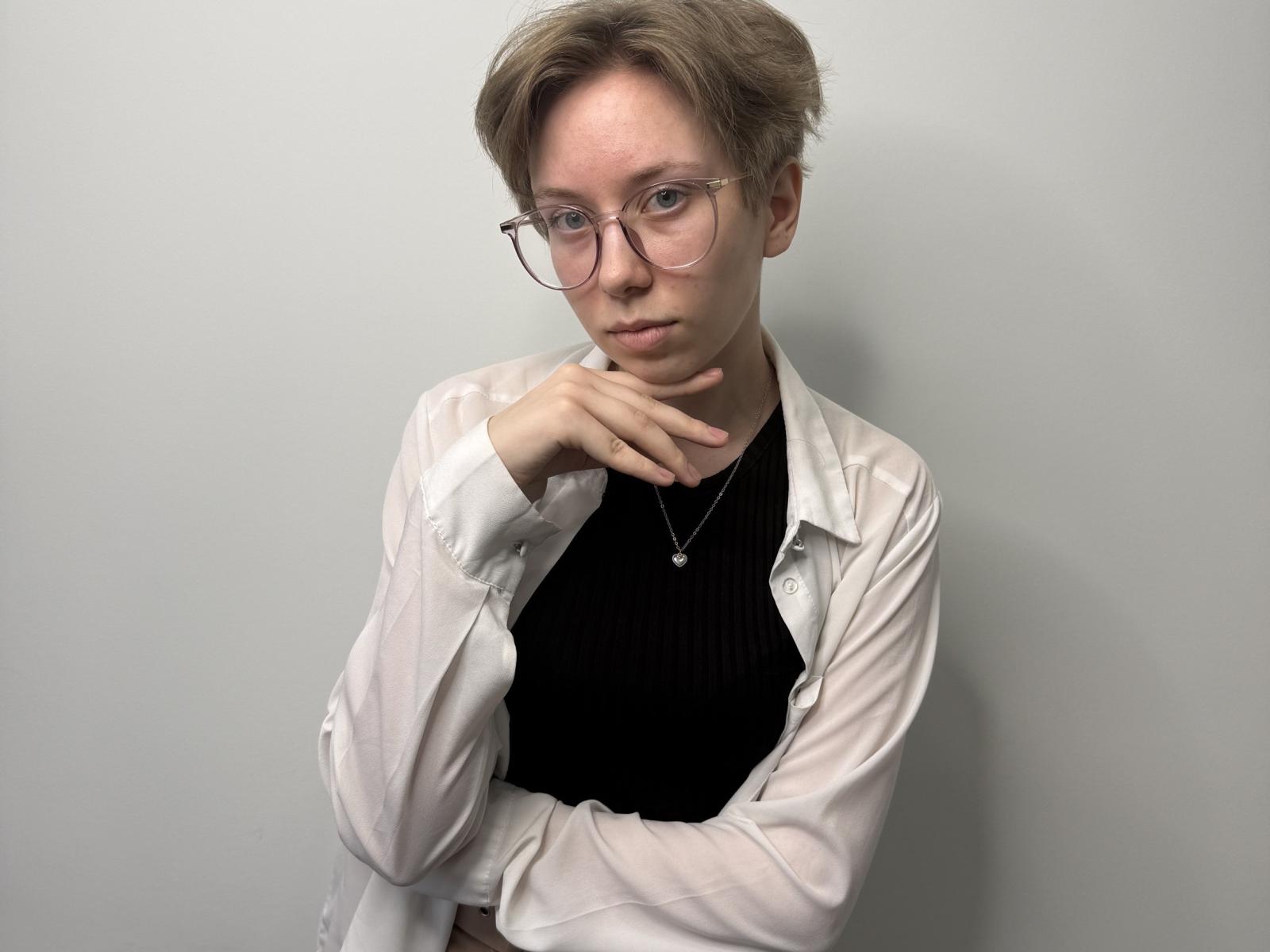 Palmira Prunier mit kurzen, blonden Haaren, Brille, weißem Hemd, schwarzem Rippstrickoberteil, Halskette, Hand am Kinn, neutraler Gesichtsausdruck, steht vor einer schlichten, grauen Wand und blickt leicht nach unten..