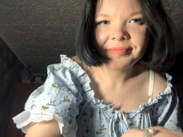 Mikaela Etherton, con cabello corto y negro, sonriendo, vestida con una blusa floral azul claro con un hombro descubierto, con luz solar en su rostro, con una correa de sostén blanca visible, piel ligeramente brillante..
