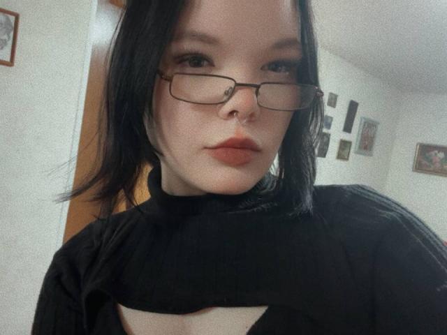 MikaelaEtherton with black hair, glasses, and black turtleneck, showing a cutout chest, has a nose ring and slightly parted lips, looking at the camera.