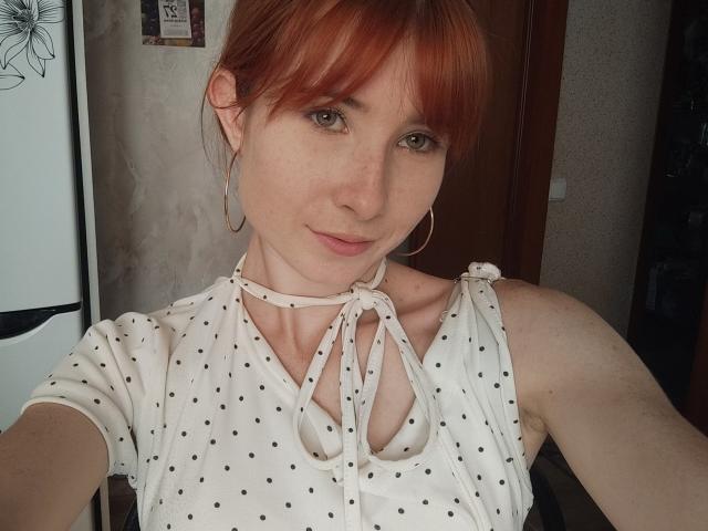 KasandraGrothe with red hair, small tits, wearing a white polka dot top with a ribbon neckline, gold hoop earrings, and a subtle smile. She's taking a selfie.