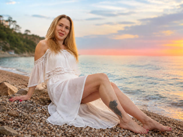 Tessa, mit langen blonden Haaren, in einem weißen, schulterfreien Kleid, sitzt auf einem felsigen Strand, die Beine gekreuzt, mit einem sichtbaren Tattoo an der rechten Wade, im Hintergrund ein Sonnenuntergang, lächelnd, barfuß, mit rosafarbenen Nägeln..