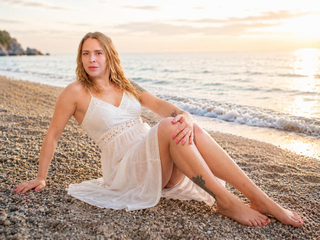 Tessa, mit welligem, blondem Haar, in einem weißen Kleid, sitzt auf einem Kiesstrand, die Beine gekreuzt, die Hände auf dem Boden, mit rosafarbenen Nägeln, einem Tattoo am rechten Oberschenkel, im Hintergrund ein Sonnenuntergang..