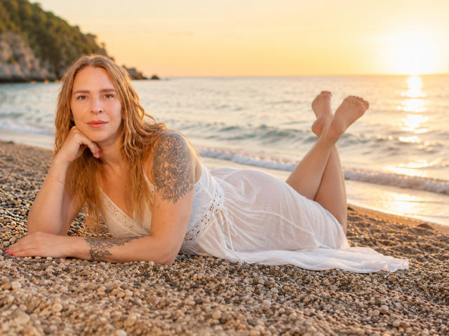 TessaDaybreak ligt op een kiezelstrand, met haar benen omhoog, gekleed in een witte, doorschijnende jurk, waarbij een tatoeage op haar rechter schouder zichtbaar is, met een ontspannen, sexy en schattige uitdrukking..