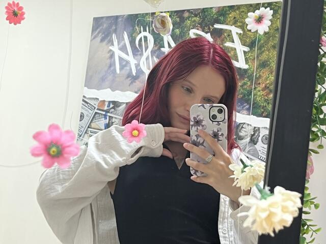 JuliannaMoor with red hair, black top, white jacket, holding phone, showing tits, mirror selfie, pink flowers, white wall, green plant, ring on finger, smiling.