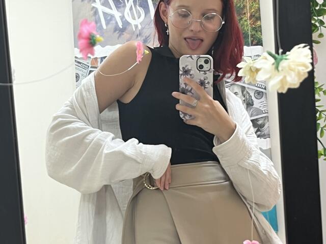 JuliannaMoor with red hair, glasses, tongue out, wearing black sleeveless top, beige skirt, white jacket, taking selfie in mirror, holding phone with floral case, showing ass.