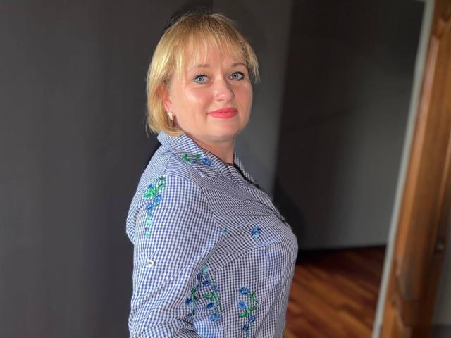 KiraSmiling has blonde hair, blue eyes, and is wearing a blue checkered shirt with floral patterns, smiling softly with red lipstick, standing in a room with wooden floors.