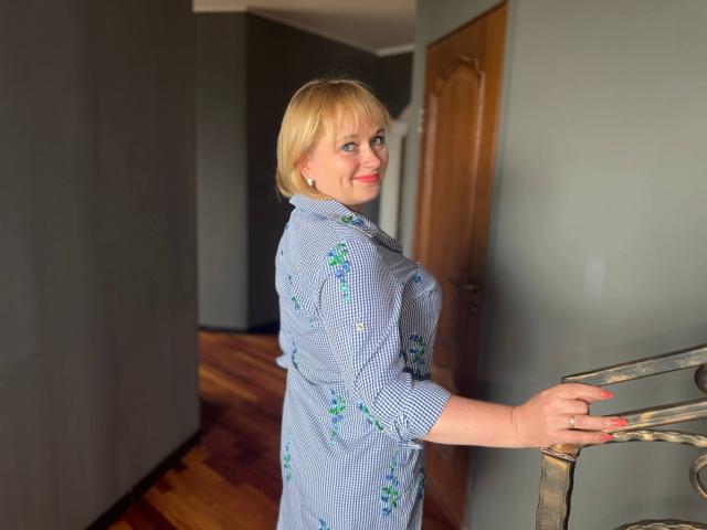 Blonde KiraSmiling with short hair, blue checkered dress with green floral patterns, standing on wooden stairs, smiling at camera, pink nail polish, holding metal railing, gray walls.