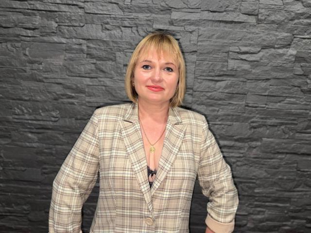 KiraSmiling has blonde hair, a plaid shirt, and a necklace, standing against a dark stone wall, with a slight smile and red lipstick, showing her tits.