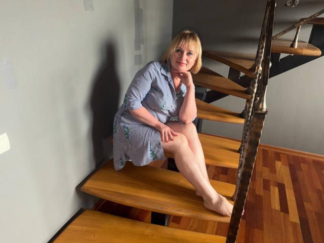 KiraSmiling sits on wooden stairs, wearing a blue floral dress, showing her ass, with one leg crossed, and her hand on her chin, barefoot, looking at the camera.